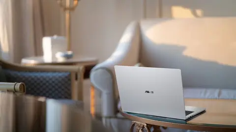 Notebook prateado da ASUS visto de trás, com o logo ASUS na tampa, sobre uma mesa de vidro em sala clara com sofá
