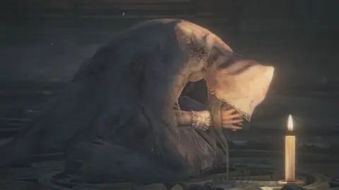 Cena de Bloodborne: figura encapuzada curvada sobre si mesma ao lado de uma vela acesa, em ambiente escuro