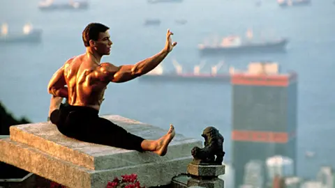 Jean-Claude Van Damme sentado em terraço com braço estendido, porto de Hong Kong ao fundo, em cena de Bloodsport