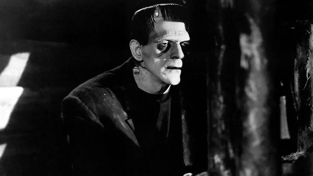 Cena em preto e branco do Monstro de Frankenstein, com parafusos no pescoço, de perfil num ambiente escuro