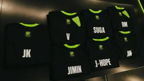 Camisetas pretas da LOUD com os nomes de membros do BTS (JK, V, Suga, RM, Jimin, J-Hope, Jin) em parede escura.