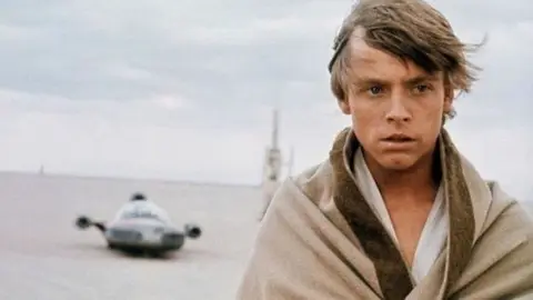 Luke Skywalker com manto bege ao lado de um landspeeder em um deserto, cena de Star Wars: Uma Nova Esperança