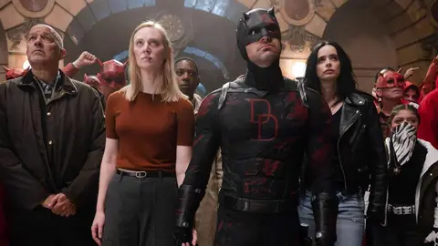 Cena da série Demolidor: homem de uniforme preto com o símbolo DD, ao lado de Krysten Ritter e outros personagens em um corredor antigo