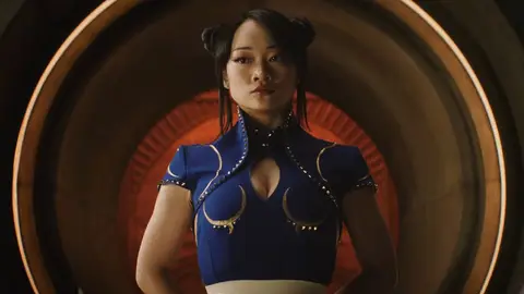 Atriz de cabelo preso em coques, com vestido azul e detalhes dourados inspirado na Chun-Li, em cenário circular laranja.