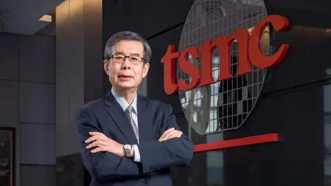 Homem de terno escuro e óculos, de braços cruzados, diante do logotipo vermelho da TSMC e de um disco de wafer