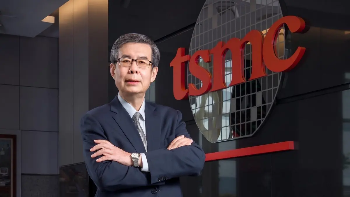 Homem de terno escuro e óculos, de braços cruzados, diante do logotipo vermelho da TSMC e de um disco de wafer