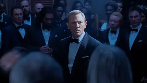 Daniel Craig de smoking e gravata-borboleta em cena de filme, cercado por convidados de traje formal em salão escuro