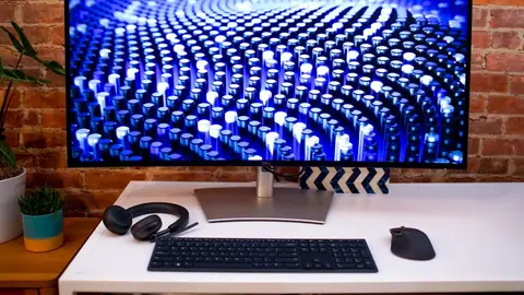 Monitor Dell com tela azul em padrão de pontos luminosos, sobre mesa branca com teclado, mouse e headset, diante de parede de tijolos