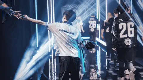 Jogadores de LoL entram em palco com luzes azuis; o da frente, de camisa branca com SHOWMAKER, estende o braço