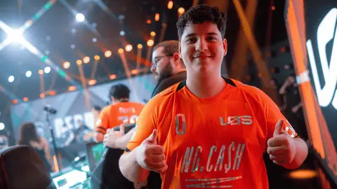 Jogador da LOS com camisa laranja da equipe NG.CASH, sorrindo e fazendo joinha em palco de evento de esports