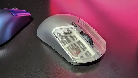 Mouse gamer cinza com laterais translúcidas sobre mesa escura, com luz rosa ao fundo