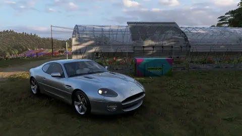Carro prateado Aston Martin parado em um gramado ao lado de estufas, com um baú rosa e verde com o logo Horizon no jogo Forza Horizon 6