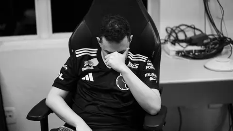 Jogador de camisa preta com patrocinadores, cabeça baixa e mão no rosto, sentado em cadeira gamer, em foto preto e branco.