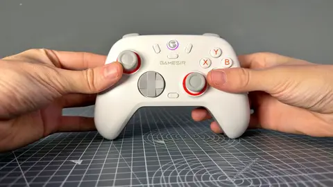 Duas mãos seguram um controle GameSir branco com detalhes vermelhos sobre um tapete de corte cinza