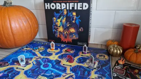 Caixa e tabuleiro do jogo de tabuleiro Horrified sobre a mesa, com abóbora e velas de Halloween ao lado