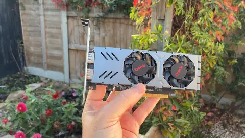 Mão segurando uma placa de vídeo NVIDIA de dois coolers na frente de uma cerca de madeira e plantas