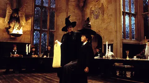 Harry Potter com o Chapéu Seletor na cabeça e a professora McGonagall ao lado, no Salão Principal de Hogwarts