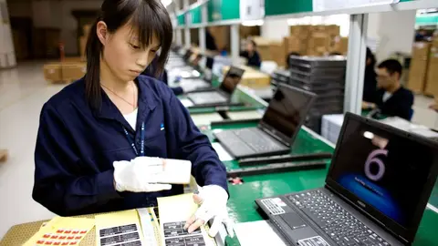 Operária de uniforme azul monta notebooks em linha de produção, com etiquetas e caixas ao fundo