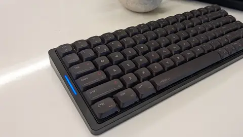 Teclado preto de baixo perfil com teclas de tampa baixa e LED azul na lateral, sobre mesa branca