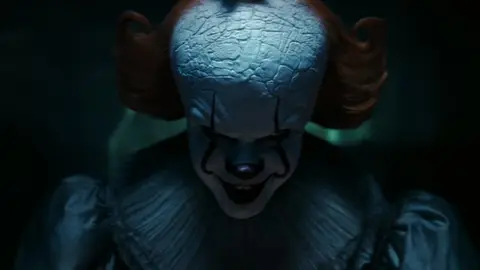 Pennywise, o palhaço de It, com maquiagem branca, cabelo ruivo e sorriso aberto em cena escura.