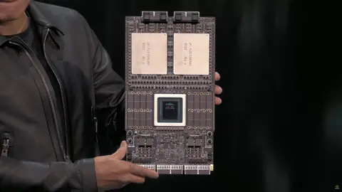Homem de jaqueta de couro segura uma placa de GPU com dois chips grandes e um chip central, em palco escuro
