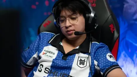 Jogador de esports de óculos e headset, com camisa azul e branca de time, sentado em cadeira gamer em evento