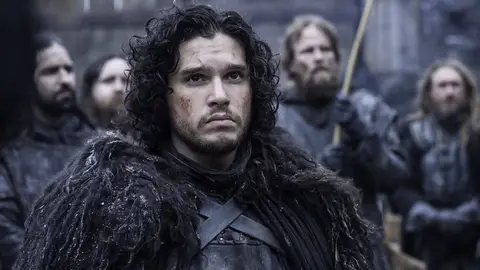 Jon Snow com capa de pele escura e homens barbudos ao fundo, em cena de Game of Thrones