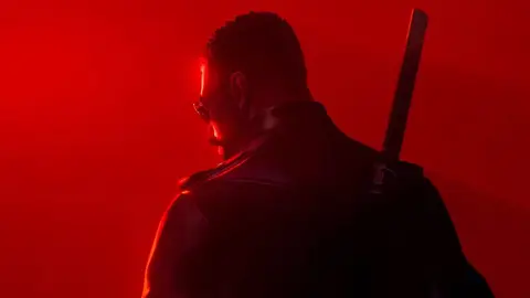 Personagem Blade de costas, com óculos escuros, jaqueta de couro e espada nas costas, sob luz vermelha intensa.