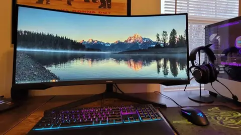 Monitor Samsung curvo em mesa de setup gamer, com teclado, mouse e headset, e paisagem de montanhas na tela