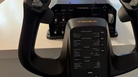 Volante de avião MOZA com painel de checklist impresso, sobre mesa clara, e módulo de painel MOZA ao fundo