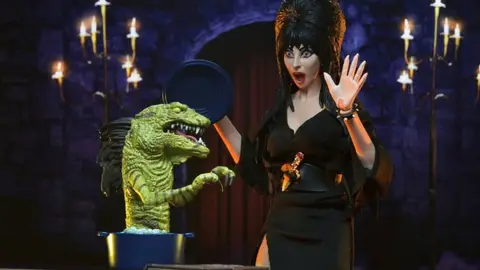 Figure da Elvira, Mistress of the Dark, ao lado de uma criatura verde, em cenário escuro com candelabros