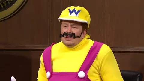 Pessoa fantasiada de Wario, com boné amarelo com W, bigode preto e macacão roxo, em cenário de programa de TV