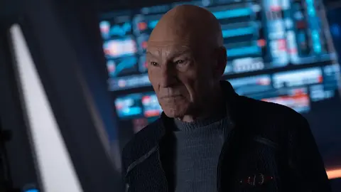 Patrick Stewart como Picard em cena de Star Trek, de uniforme escuro, em ponte de comando com painéis azuis ao fundo