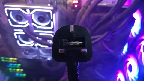 Plugue britânico de 13 A com fusível queimado e enegrecido, em primeiro plano, com um PC iluminado por LEDs RGB ao fundo
