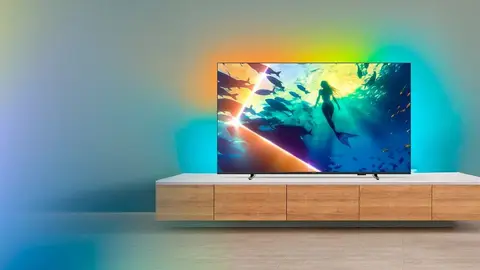 TV Philips Ambilight ligada sobre móvel de madeira, com luz colorida projetada na parede e cena de sereia na tela