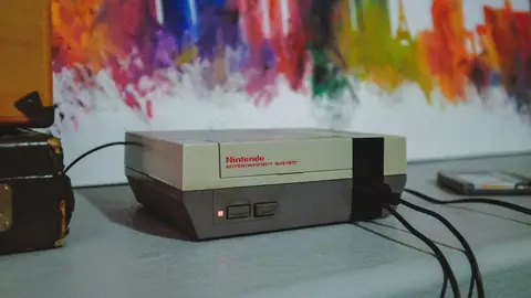 Console Nintendo Entertainment System (NES) sobre uma mesa, com fios conectados e painel colorido ao fundo