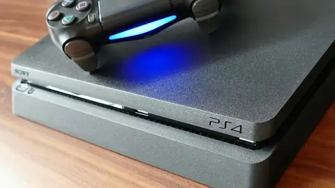 Console PS4 Slim com controle DualShock 4 e luz azul acesa, sobre mesa de madeira
