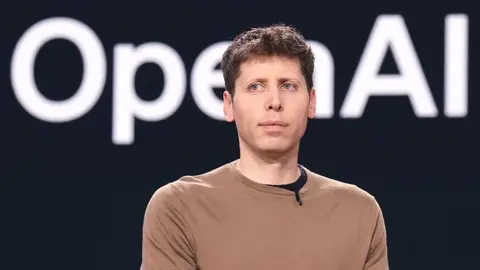 Sam Altman, de suéter marrom, em frente a um telão escuro com a palavra OpenAI em letras brancas