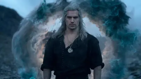 Geralt de Rívia, de cabelo grisalho e medalhão do lobo, diante de um portal azulado em cena de The Witcher