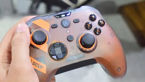 Mão segurando controle SCUF translúcido laranja, com botões ABXY e analógicos em destaque