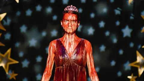 Personagem de Carrie coberta de sangue falso, com tiara, vestido vermelho e fundo de estrelas douradas e azuis