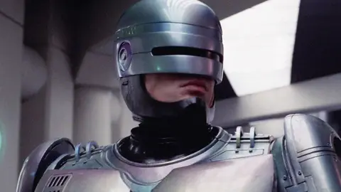 Cena do RoboCop original, com o policial ciborgue de armadura prateada e visor escuro, em ambiente interno