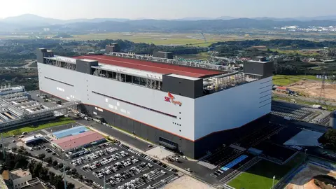 Vista aérea de grande fábrica da SK hynix, com telhado vermelho e logo da empresa na fachada, cercada por estacionamento