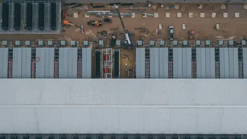 Vista aérea de canteiro de obras de data center, com fileiras de módulos técnicos e galpão branco de grande porte