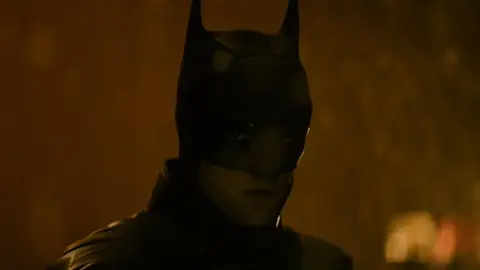 Batman de Robert Pattinson, com máscara e capa, em cena escura e alaranjada, olhando para a câmera