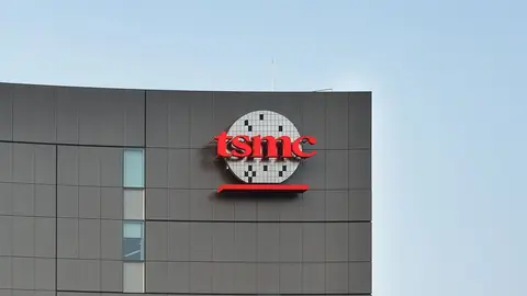 Fachada de prédio cinza com o logo vermelho da TSMC sobre um disco de wafer de silício, sob céu azul claro