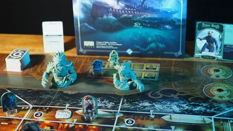 Tabuleiro do jogo Unfathomable montado sobre uma mesa de madeira, com miniaturas azuis de monstros, peças e a caixa do jogo ao fundo