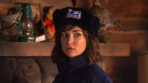 Mulher de gorro de pele com emblema de correio e jaqueta azul-escura, olhando para o lado, em cabana com lanterna e aves empalhadas