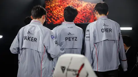 Jogadores da T1 de costas com jaquetas da equipe, nomes Taker, Peyz e Oner, diante de telão em arena de LoL